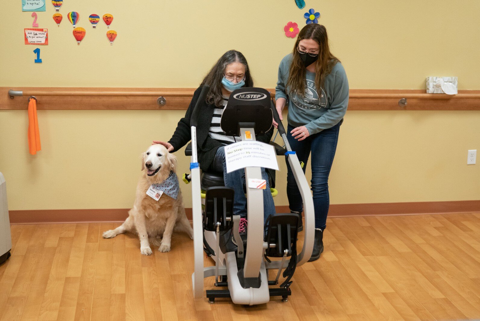A therapy dog and a staff member help a participant on an exercise bike.